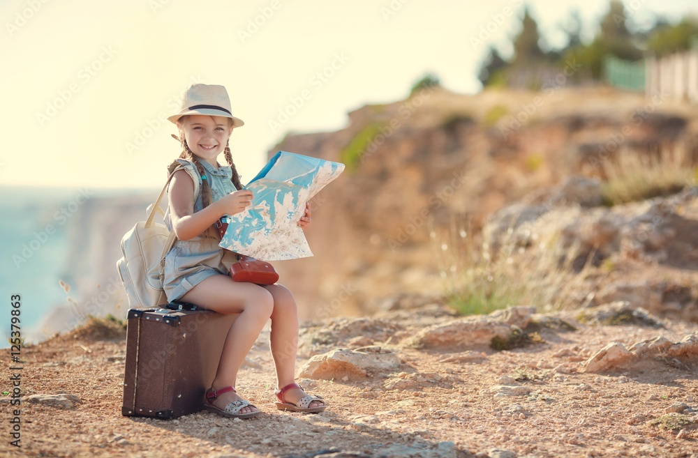 Cute little girl,long hair braided into two pigtails,on his head wearing a sun hat,behind wears light leather backpack,sitting on the old suitcase among the back roads,exploring the map of the area