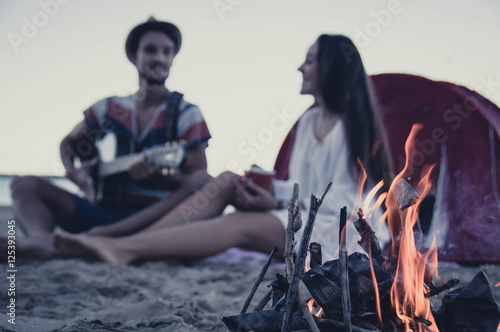 Fototapeta Happy young couple sitting near campfire and playing guitar