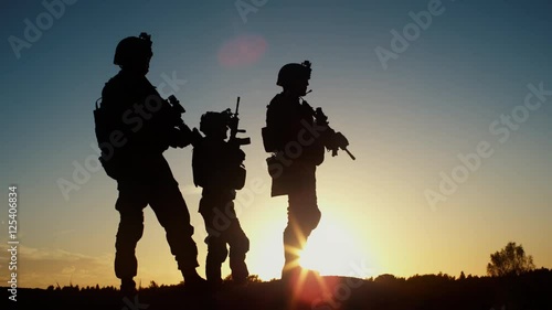 Squad of Three Fully Equipped and Armed Soldiers Standing in Desert Environment in Sunset Light. Slow Motion. photo