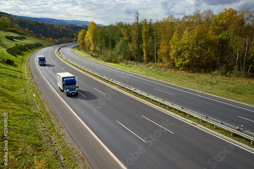 Asphalt highway with oncoming blue small trucks in the autumn landscape.