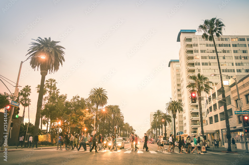 Naklejka premium Mieszkańcy i turyści spacerujący po przejściu dla pieszych i po Ocean Ave w Santa Monica po zachodzie słońca - Zatłoczone ulice stanu Los Angeles i Kalifornia - Ciepłe, desatowe odcienie zmierzchu z zamazanymi ludźmi