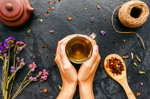 Woman's hands holding cup of tea on black background with rustic dried flowers, herbs. Top view, Flat lay.