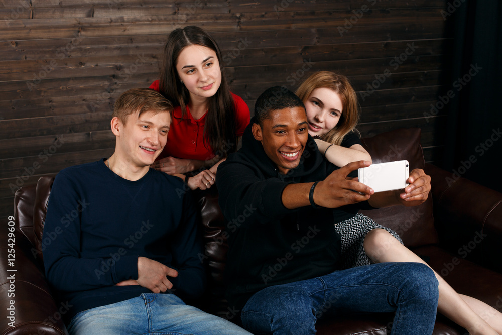 Friends making joint portrait using camera in phone. Four friends make selfie sitting on the couch. Multicultural friends