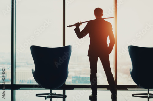 Young businessman in office in business suit standing between of two armchairs in front of window and holding katana sword like japanese samurai warrior, blurred winter city top view in background photo