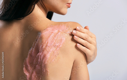 Young woman applying scrub on shoulder on grey background