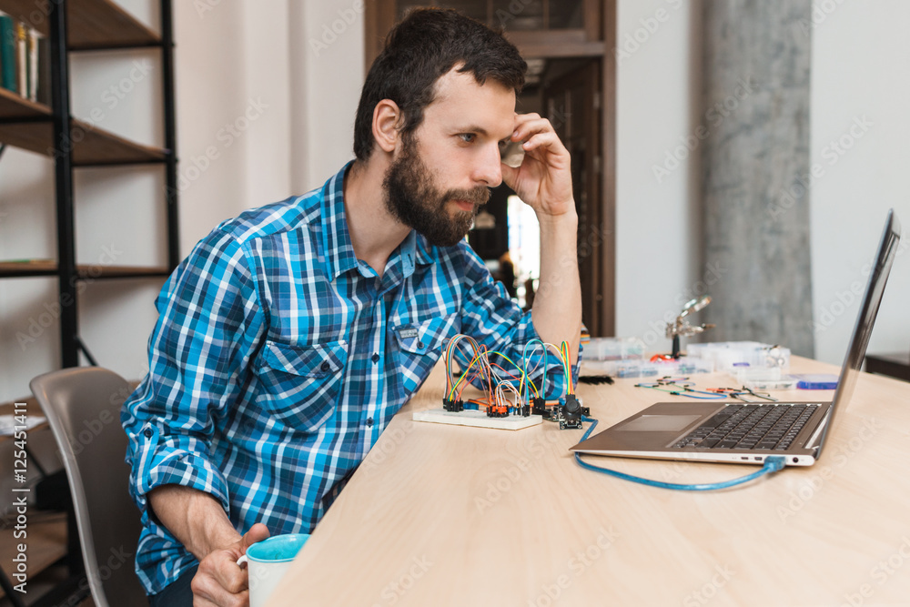 © golubovy - Bearded engineer waiting for program download, free space. Programmer sitting at workplace with cup of coffee and looking at laptop screen, testing new computer program