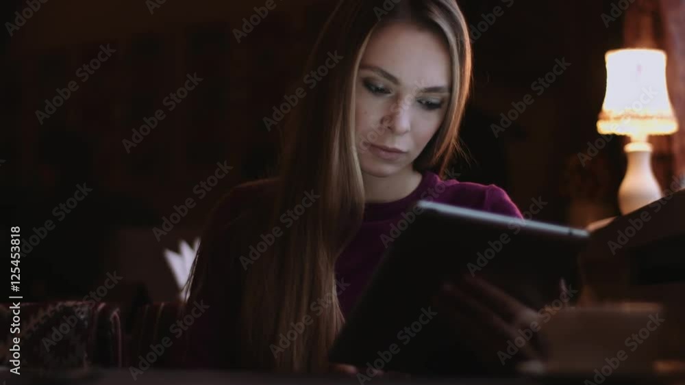 Beautiful woman with long hair uses tablet computer in the cafe, her face illuminated by the screen Beautiful woman with long hair uses tablet computer in the cafe, her face illuminated by the screen