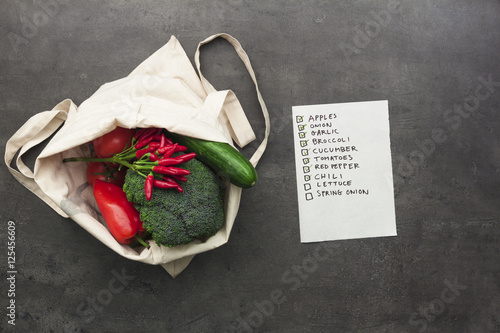 Cotton shopping bag full of vegetables and fruits with checked shopping list. Flat lay food on table.