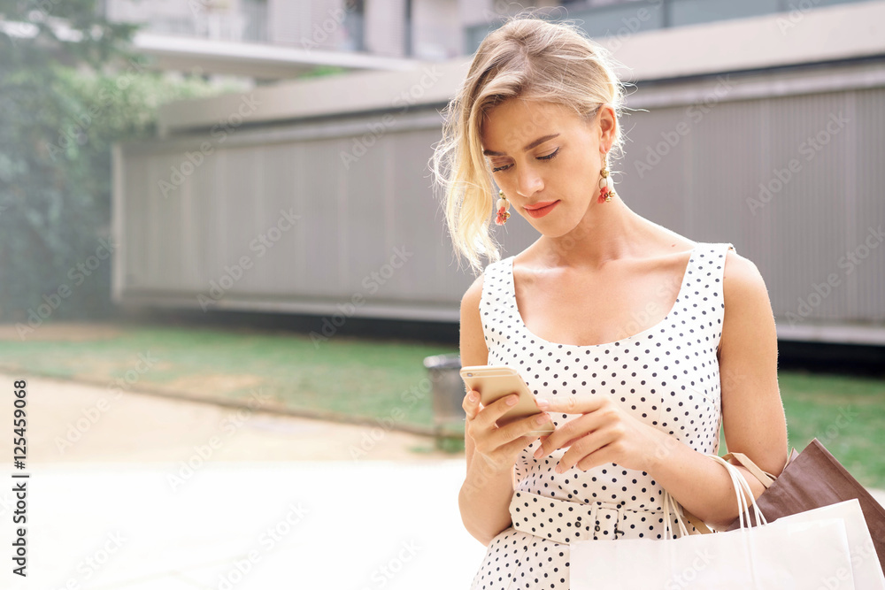 A waist up photo of a charming blondy hipster girl wearing white summer dress with black polka dots and standing outdoors on a sunny day with a paper bags and a smartphone in the hands.Online shopping