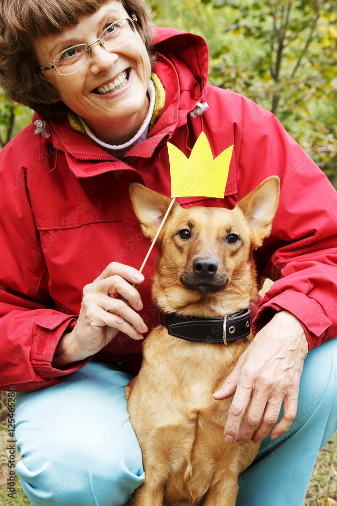Old, smiling woman holding paper crown on stick close to dog's muzzle. Dog in funny, fake crown with ownerin park. Old, smiling woman holding paper crown on stick close to dog's muzzle. Dog in funny, fake crown with ownerin park.