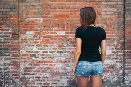Obraz na plátně An outdoor portrait of a young pretty hipster girl with long dark hair wearing black blank t-shirt and blue jeans shorts standing with her back to the camera on the brick wall background