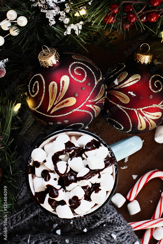 Enamel cup of hot cocoa for Christmas with mini marshmallows and garnished with chocolate sauce.  Surrounded by warm gray scarf, ornaments, pine bough and candy canes against a rustic background.