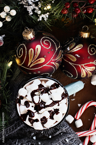 Enamel cup of hot cocoa for Christmas with mini marshmallows and garnished with chocolate sauce. Surrounded by warm gray scarf, ornaments, pine bough and candy canes against a rustic background.