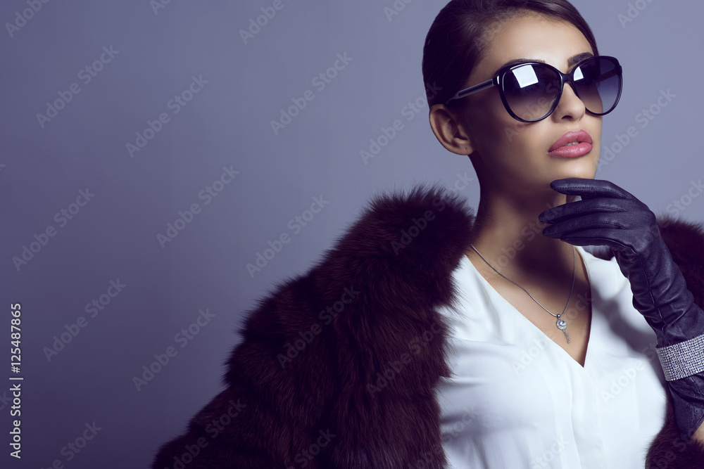 Portrait of glam dark-haired model in stylish classic sunglasses wearing white blouse, sable coat and set of luxurious bracelet and silver chain with pendant. Eyewear concept. Studio shot. Copy-space Portrait of glam dark-haired model in stylish classic sunglasses wearing white blouse, sable coat and set of luxurious bracelet and silver chain with pendant. Eyewear concept. Studio shot. Copy-space