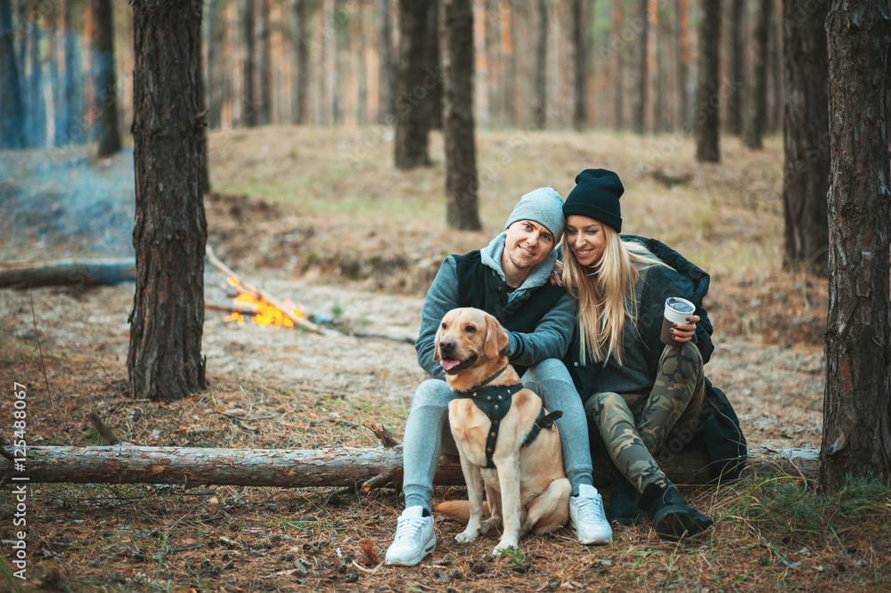 Fototapeta premium Romantic couple with dog sitting near bonfire, autumn forest background. Young blonde woman and handsome man. Concept - family, togetherness, love, friendship.