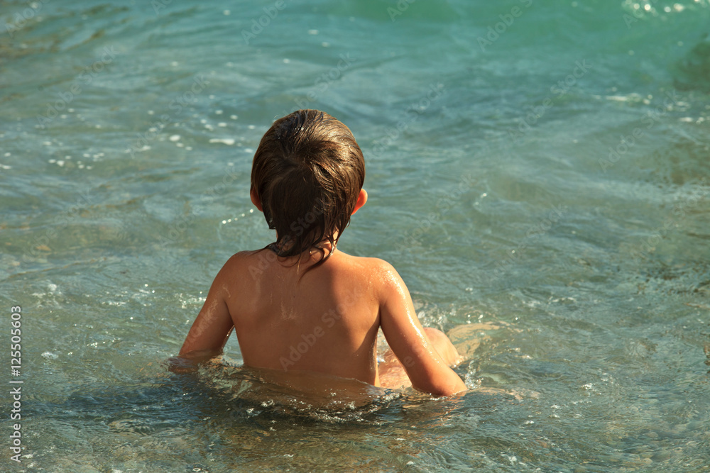 Boy of five years bathing in the sea. He is sitting in the water back to the camera