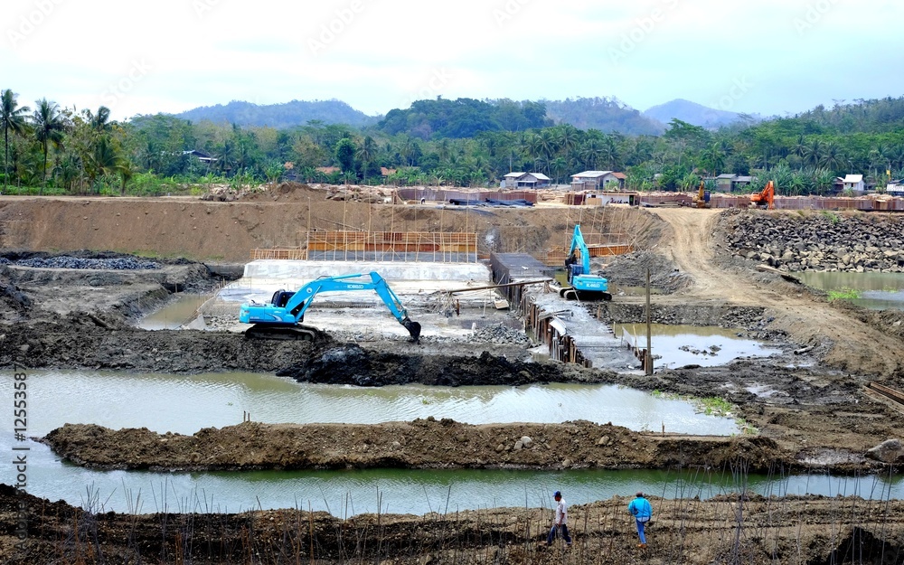 Naklejka premium Blue Excavator working in construction site and few worker appear walking in the site