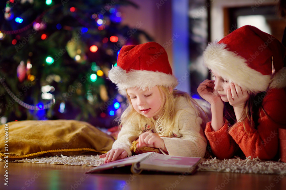 Two cute little sisters reading a story book together under a Christmas tree Two cute little sisters reading a story book together under a Christmas tree
