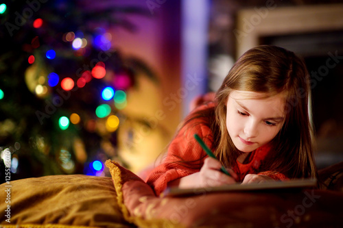 Cute little girl writing a letter to Santa by a fireplace on Christmas