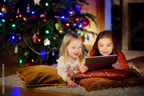 Two cute little sisters using a tablet pc by a fireplace on Christmas evening