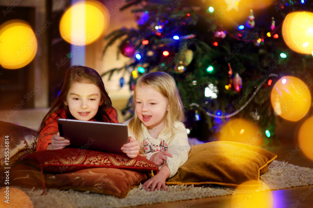 Two cute little sisters using a tablet pc by a fireplace on Christmas evening Two cute little sisters using a tablet pc by a fireplace on Christmas evening