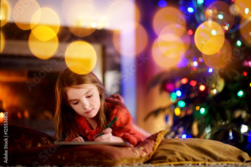 Cute little girl writing a letter to Santa by a fireplace on Christmas