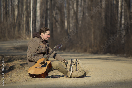 A man with a backpack and a guitar sitting by the roadside