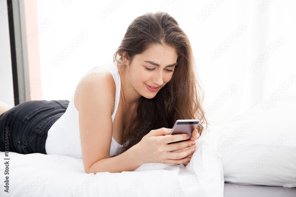 Beautiful Caucasian woman laying down on white bed using mobile phone and smile Beautiful Caucasian woman laying down on white bed using mobile phone and smile