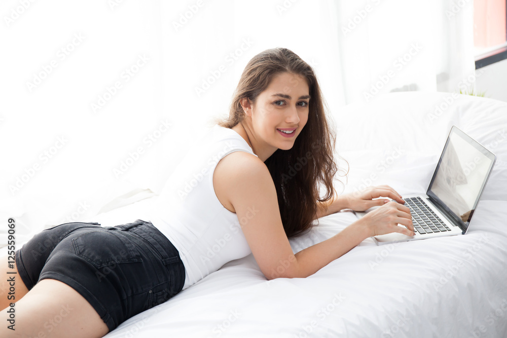 Beautiful Caucasian woman laying down on white bed using laptop and smile Beautiful Caucasian woman laying down on white bed using laptop and smile