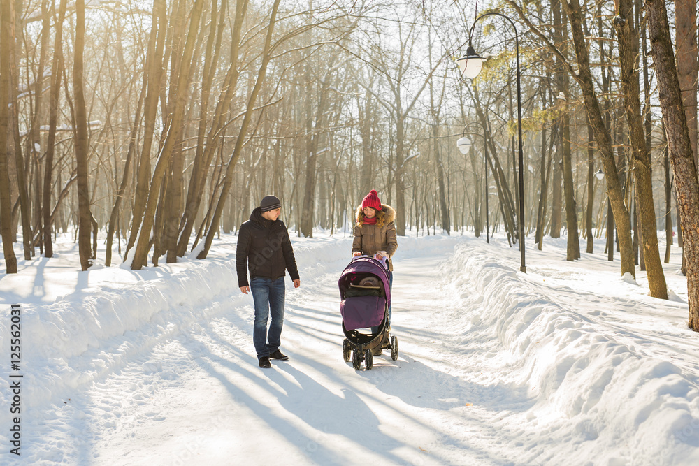 Happy young family walking in the park in winter. The parents carry the baby in a stroller through the snow.