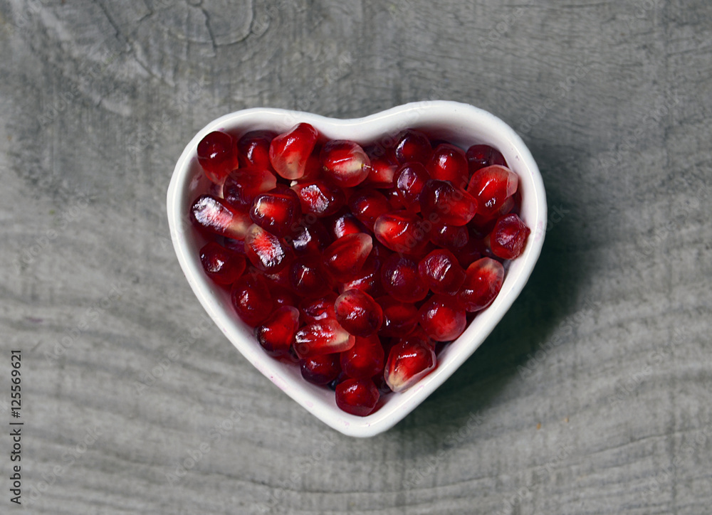 Pomegranate seeds in a white heart shaped bowl on old wooden rustic background.Healthy food or diet concept.Selective focus.Copy space. Pomegranate seeds in a white heart shaped bowl on old wooden rustic background.Healthy food or diet concept.Selective focus.Copy space.
