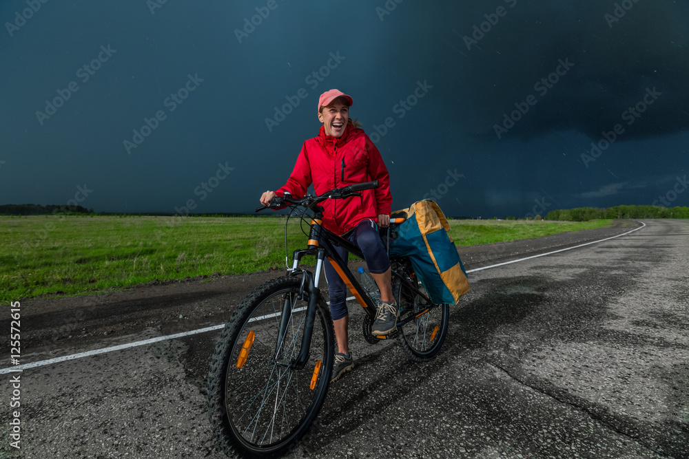 Lady hiker standing with loaded bicycle on the paved asphalt road with stormy clouds on the background
