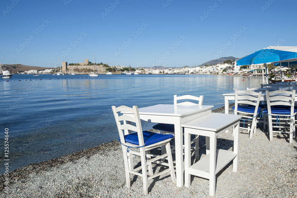 Simple beachside tables line the pebble shore of the tourist resort of Bodrum, Turkey with a scenic view of the castle