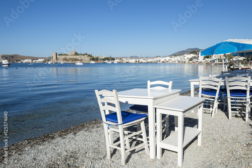 Simple beachside tables line the pebble shore of the tourist resort of Bodrum, Turkey with a scenic view of the castle