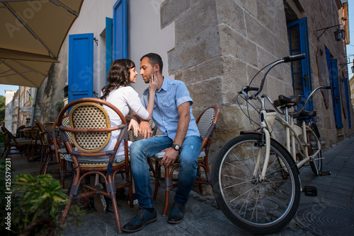 Loving couple sitting in sidewalk cafe near their tandem bike. Wife's hand gently concerns cheeks her husband, they tenderly looking at each other