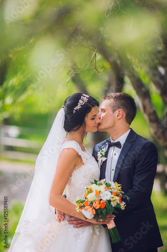 Elegant stylish groom with his happy gorgeous brunette bride on the background of trees in the park. Kissing picture.