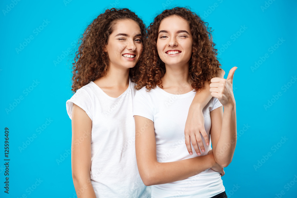 Two girls twins smiling, winking, showing like over blue background.