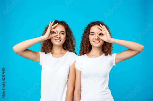 Two pretty girls twins smiling, joking over blue background.