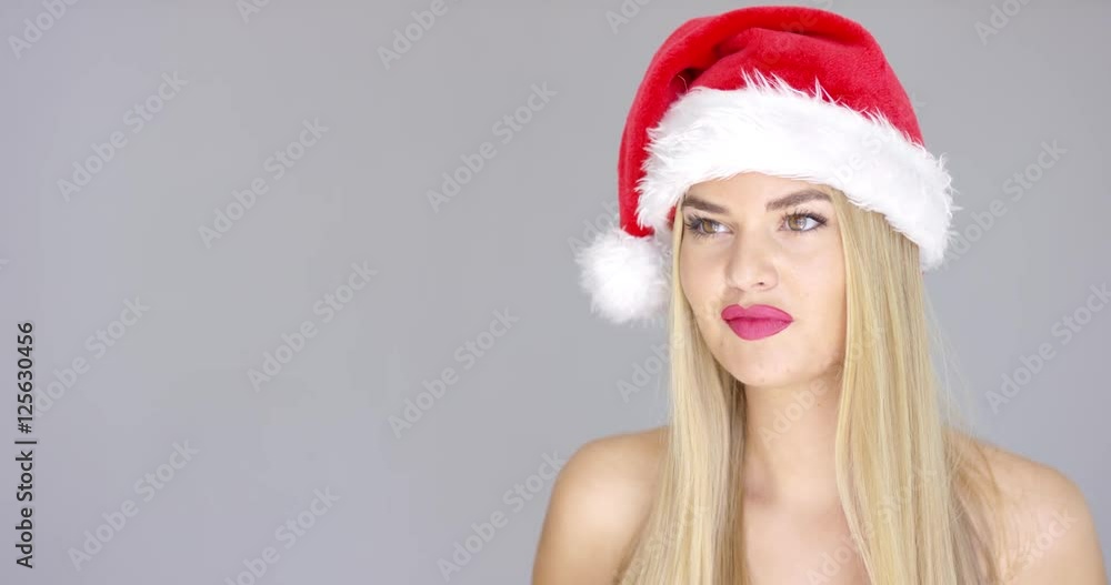 Young sexy woman in Santa Claus Christmas hat posing isolated on gray background. Smiling to the left side of the frame. She is bare shoulders. Young sexy woman in Santa Claus Christmas hat posing isolated on gray background. Smiling to the left side of the frame. She is bare shoulders.