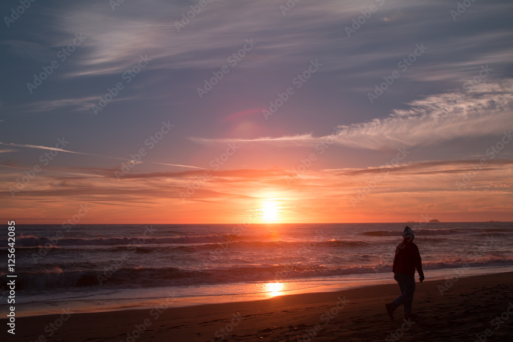A woman walks on the beach at sunset.