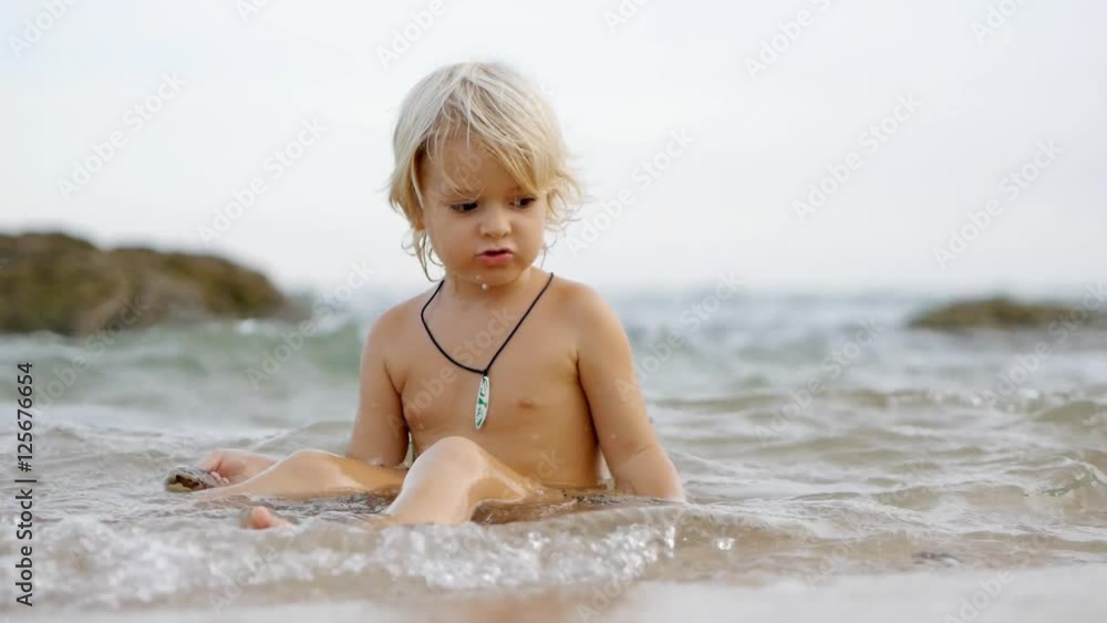 Little cute smiling child playing with shells and stones  on waves in blue sea. Having fun in summer vacation. Static view 