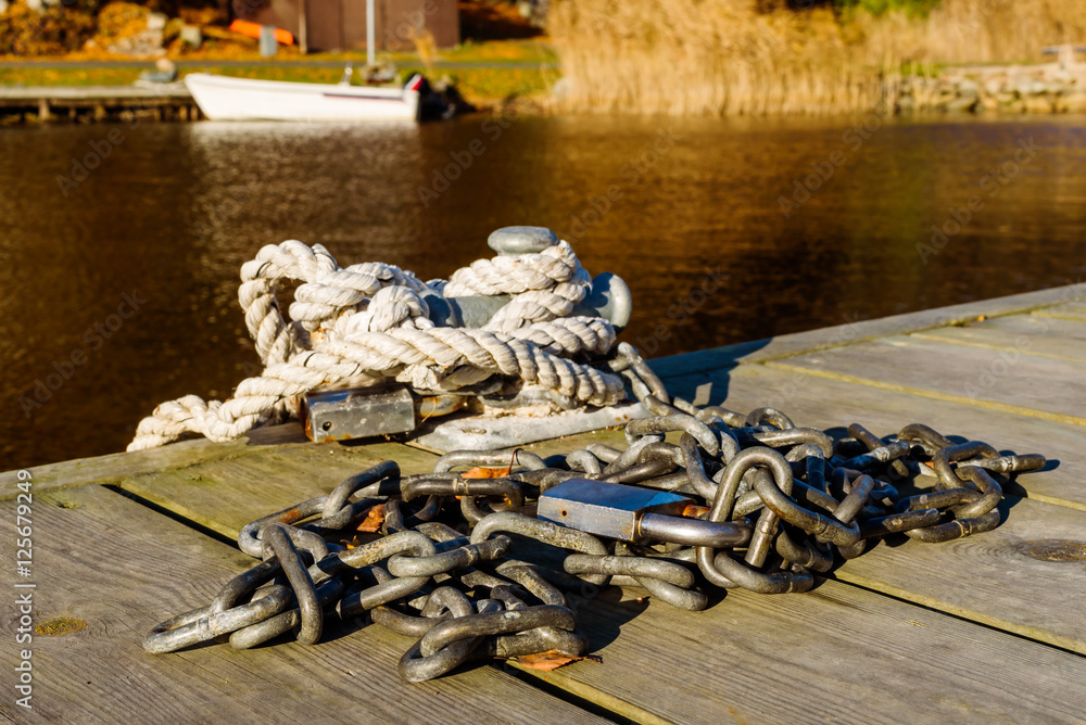 Fototapeta premium Steel chain with padlock on wooden pier. Rope and padlock on bollard. Blurred background with moored boat and coastline.