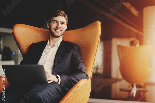 Young smiling successful man entrepreneur in formal business suite with a beard sitting on orange armchair with laptop in luxury office interior photo