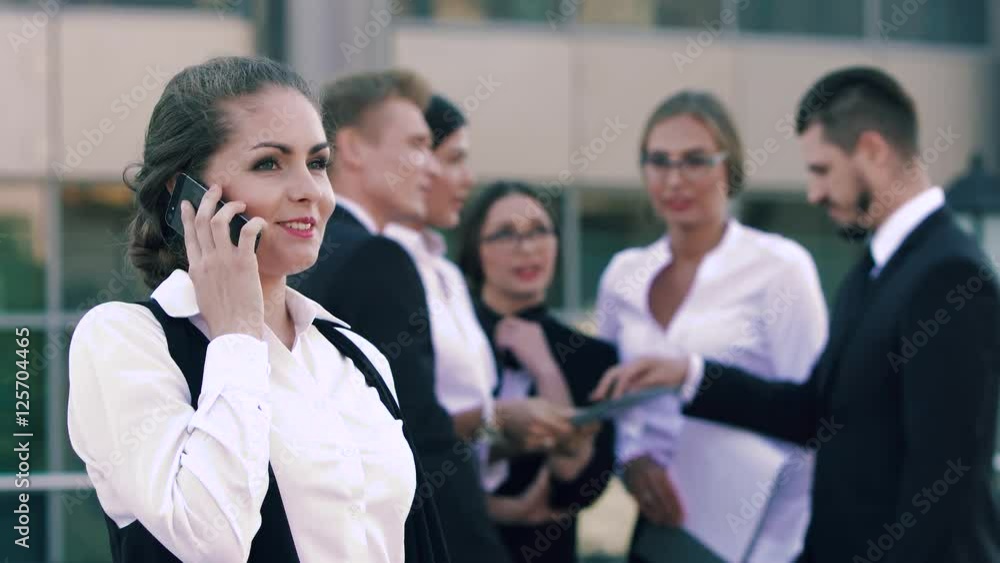 Young smiling business woman discussing her successful working day on the phone and her colleagues chatting in the background Young smiling business woman discussing her successful working day on the phone and her colleagues chatting in the background