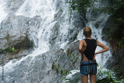 Back view of blonde woman standing on a rock and looking up near waterfall. Female tourist enjoying by a water fall in forest