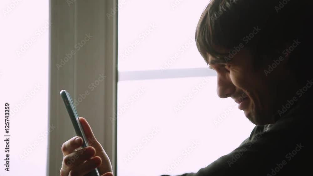Sad young man using a smart phone at the window at home in cloudy bad weather. 1920x1080 Sad young man using a smart phone at the window at home in cloudy bad weather. 1920x1080
