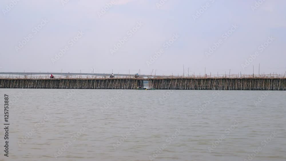 The small fishing boat is passing by bamboo bridge and motorbikes crossing it