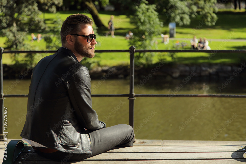 An from the angle photo of a man sitting on a bench wearing a black jacket, with a river and grass behind him. Sunny summer day. Stock | Adobe