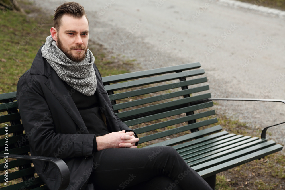 Fototapeta premium A lonely depressed man sitting down outdoors on a bench on a cold fall day wearing a black jacket, scarf and jeans.