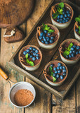 Homemade Tiramisu dessert in glasses with cinnamon, mint and fresh blueberries in wooden tray and sieve with cocoa powder over rustic wooden background, top view, selective focus, vertical composition